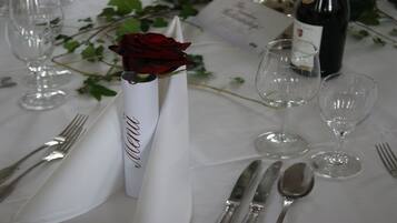 Ein Tisch ist für eine festliche Veranstaltung gedeckt, mit einer weißen Tischdecke, einer Rose in einer weißen Vase sowie Weingläsern und Besteck.