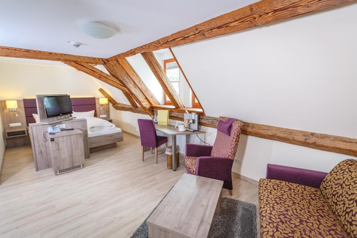 A room with a sloped ceiling, wooden beams, a bed with a TV, a desk, and purple chairs.