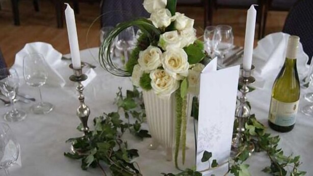 Une table dressée pour un événement formel, avec des nappes blanches, un vase de fleurs blanches, des bougies et une bouteille de vin.
