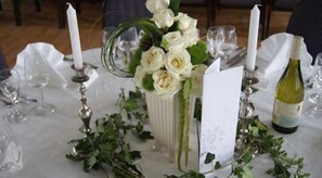 Une table dressée pour un événement formel, avec des nappes blanches, un vase de fleurs blanches, des bougies et une bouteille de vin.