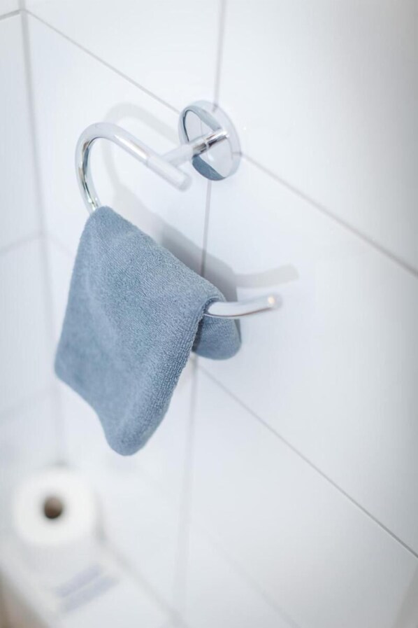 Une salle de bain avec un porte-serviettes sur lequel est posée une serviette bleue, des carreaux blancs et un rouleau de papier toilette.