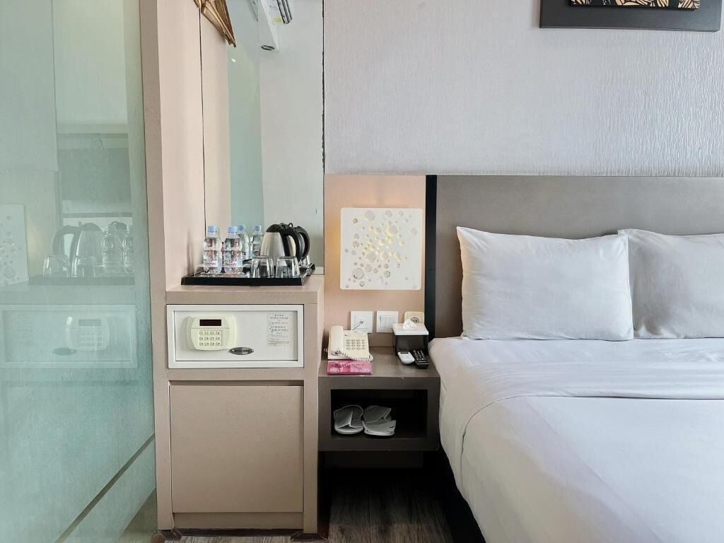 Hotelkamer met een bed, nachtkastje en een spiegel. Er is een bedieningspaneel aan de muur, een klein plankje met schoenen en een lijst met een foto.