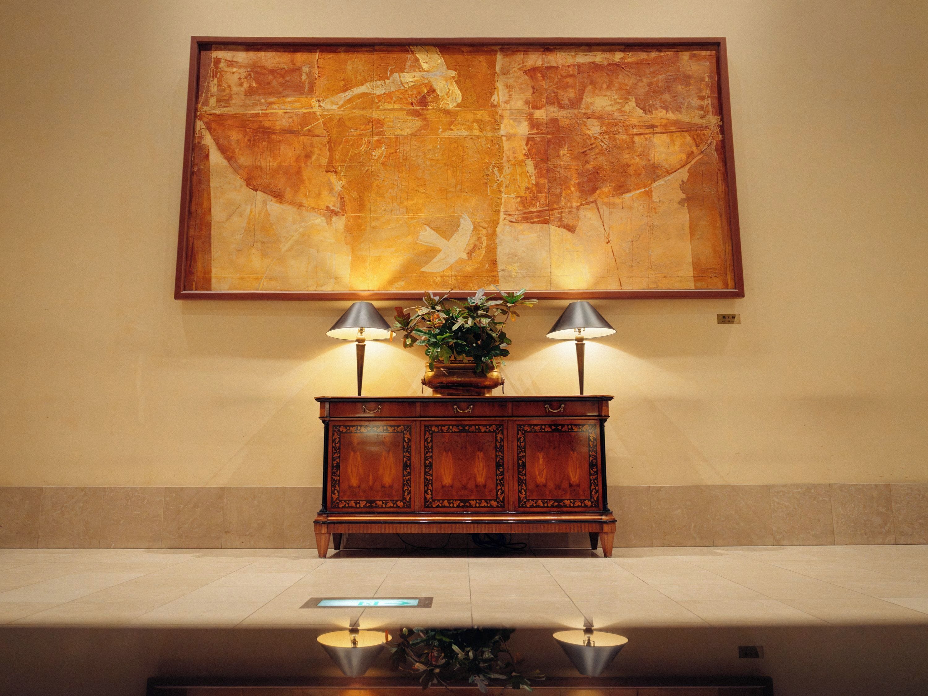 Un hall con un gran cuadro abstracto, una mesa consola de madera con dos lámparas y una planta.