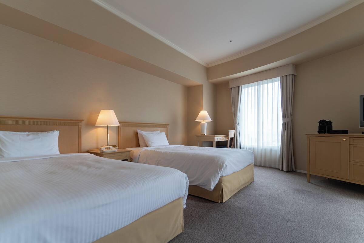 Quarto de hotel com duas camas, mesas de cabeceira com abajures, escrivaninha, cadeira e televisão sobre um móvel.
