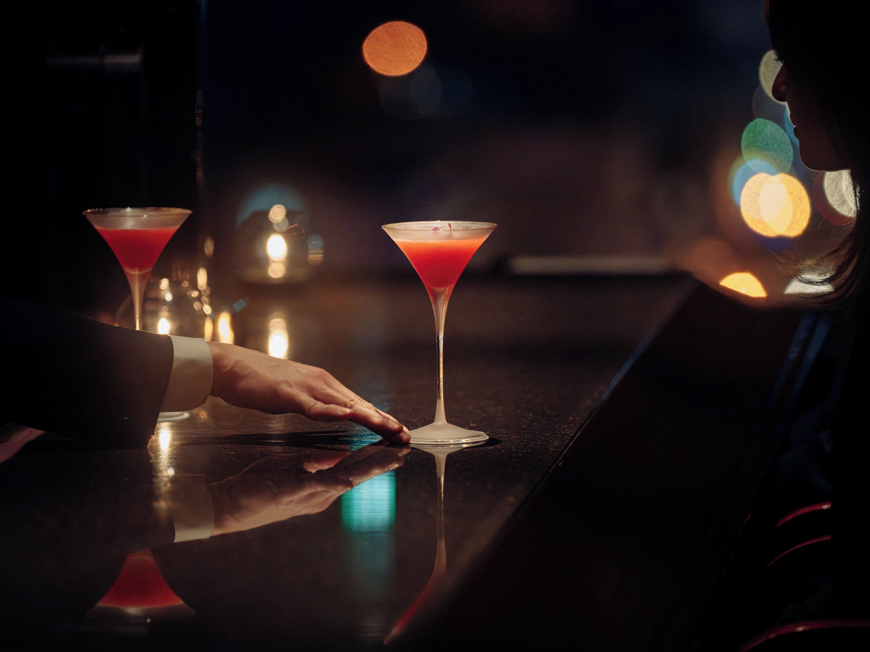 A bar setting with two martini glasses on a reflective surface, a hand reaching for one of them, and a blurred background with lights.