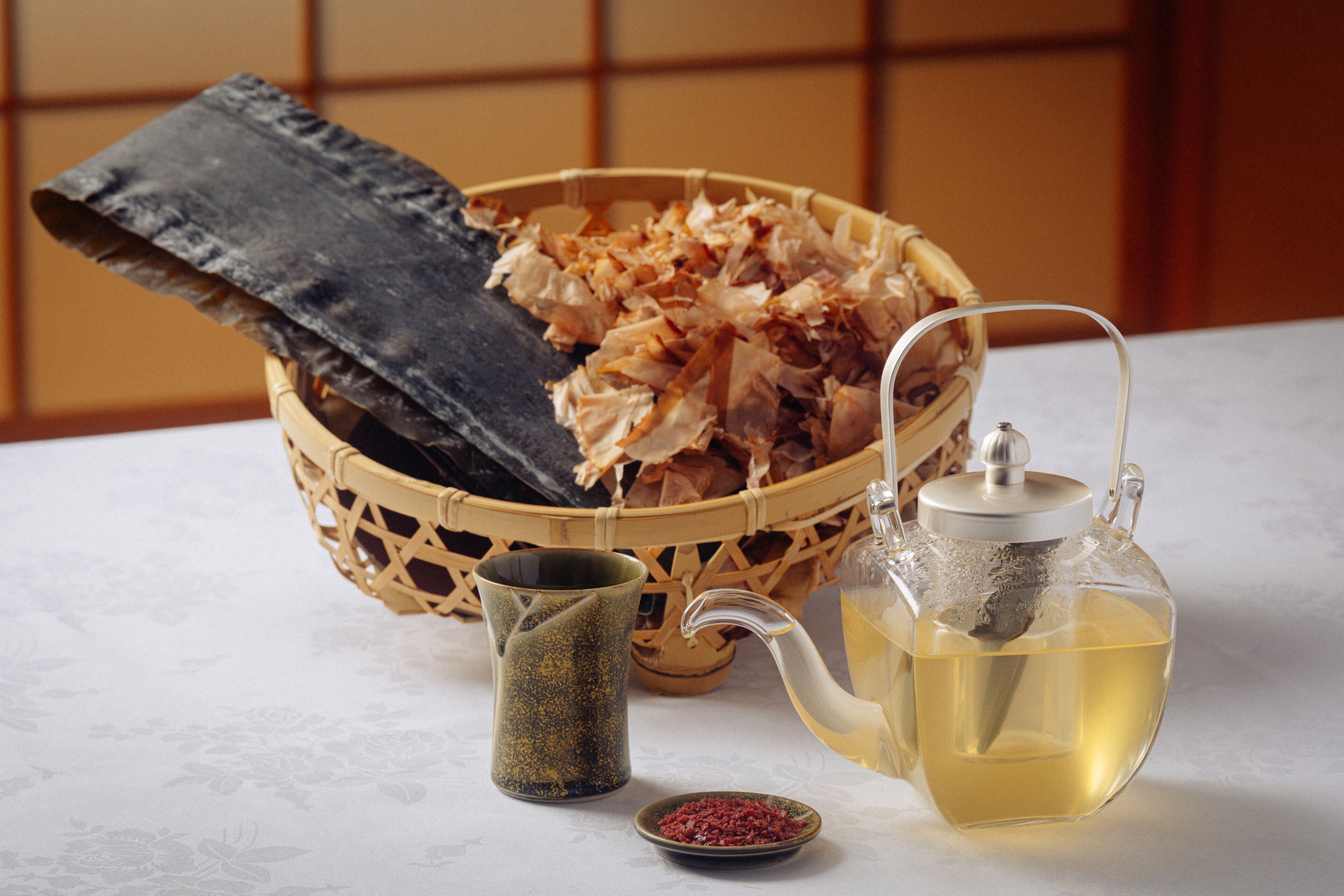 A woven basket filled with dried seaweed, a ceramic teapot with tea, and a small ceramic cup with red powder.