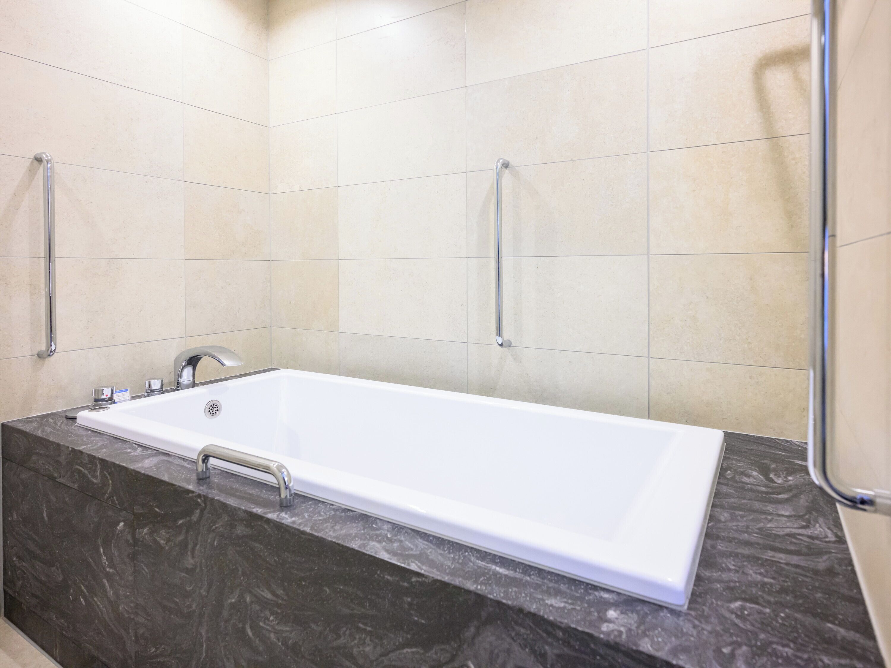 A modern bathroom with a white bathtub, dark marble countertop, and beige tiled walls.