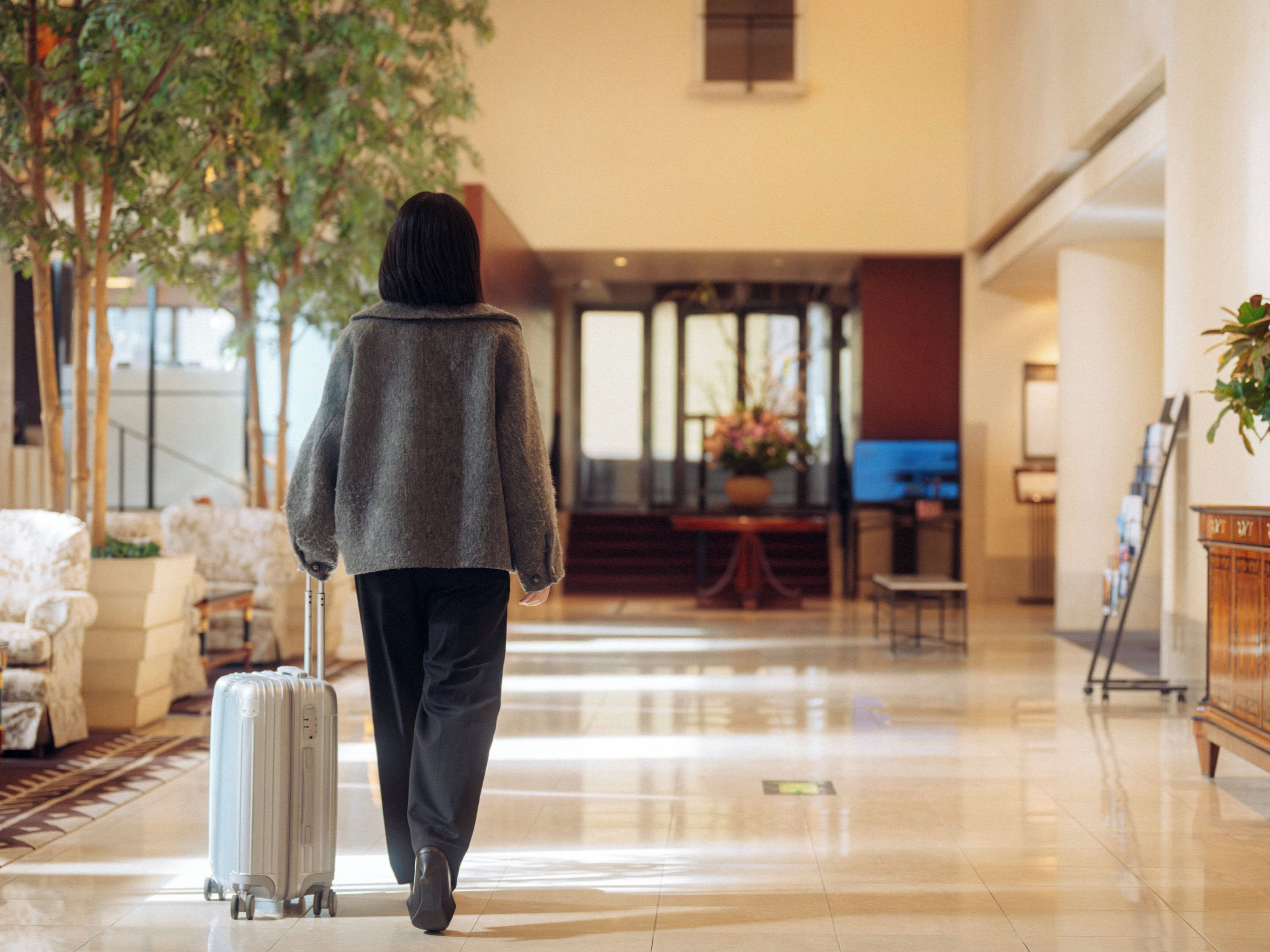 A person walking with a suitcase in a hotel lobby with a plant, a reception desk, and a seating area.
