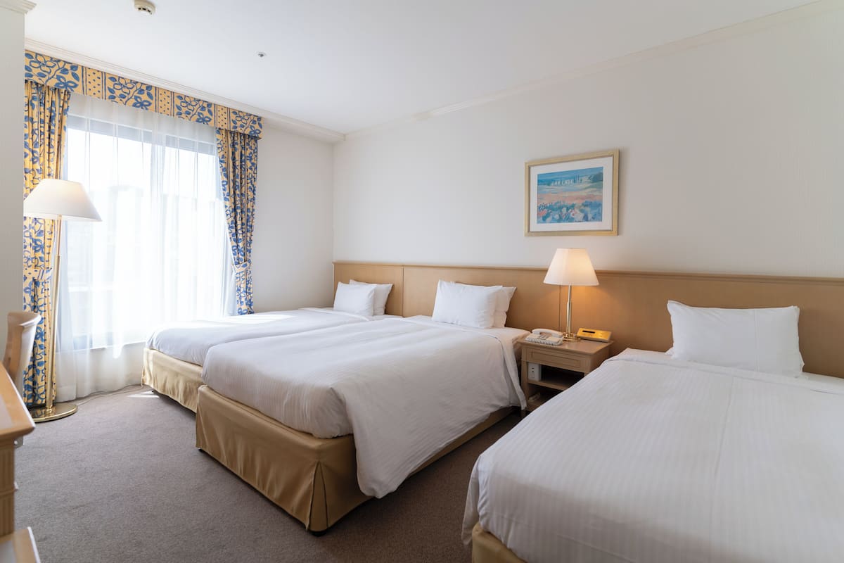 Quarto de hotel com três camas, uma mesa de cabeceira com luminária, um quadro na parede e uma janela com cortinas.