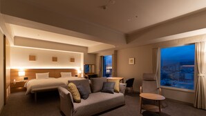 Habitación de hotel con cama, sofá, escritorio y vista a la ciudad al anochecer.