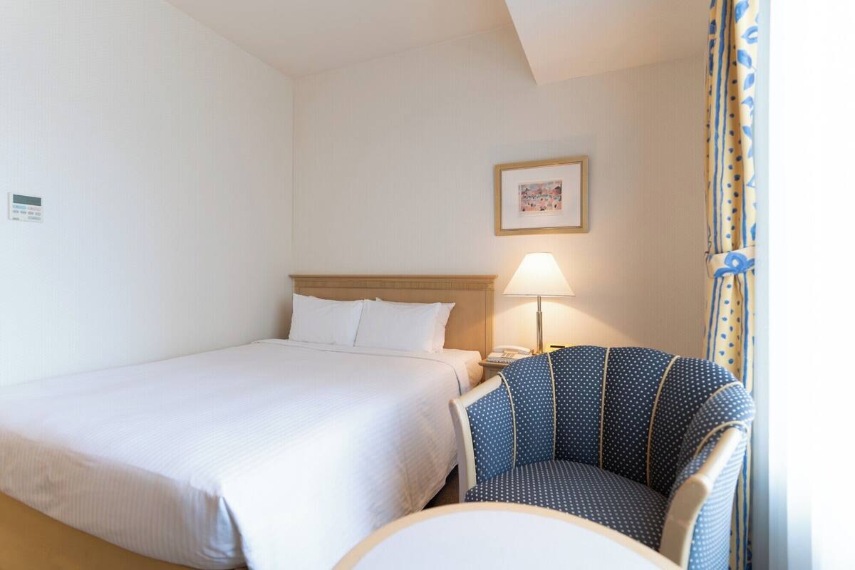 Quarto de hotel com uma cama, uma cadeira, mesa de cabeceira com abajur e quadro na parede.