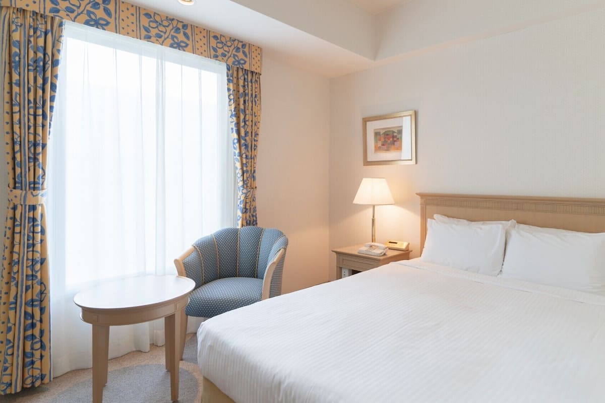 Hotellihuone, jossa on sänky, tuoli, pöytä, lamppu ja taulu seinällä.