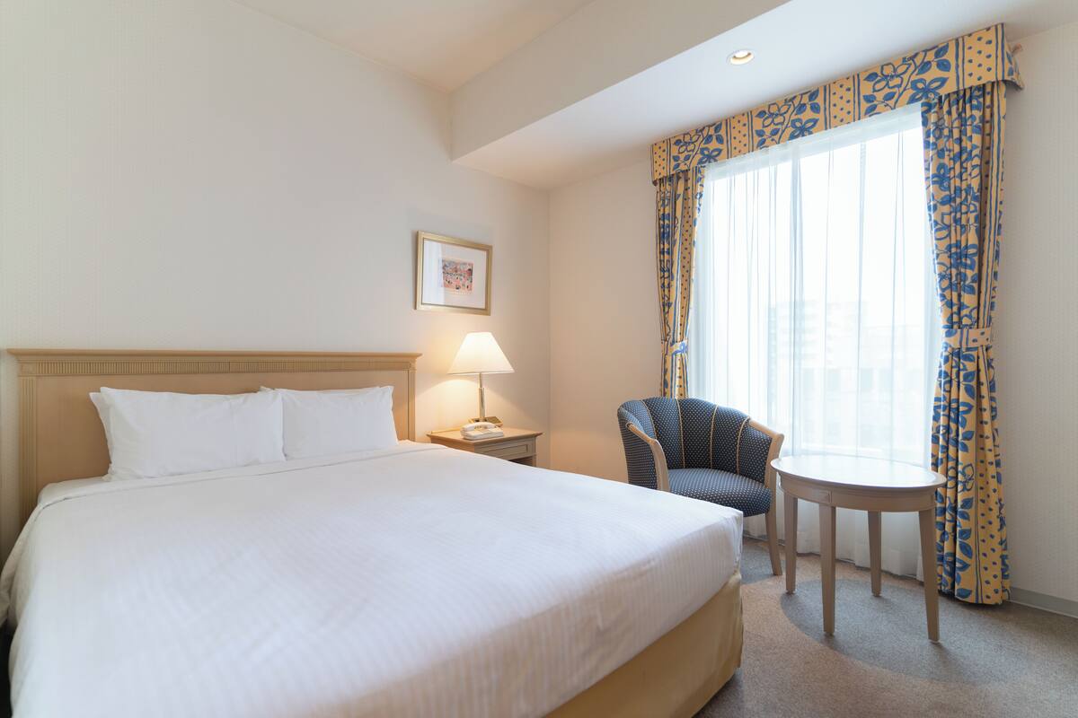 Quarto de hotel com uma cama grande, mesa de cabeceira com luminária, cadeira e janela com cortinas.