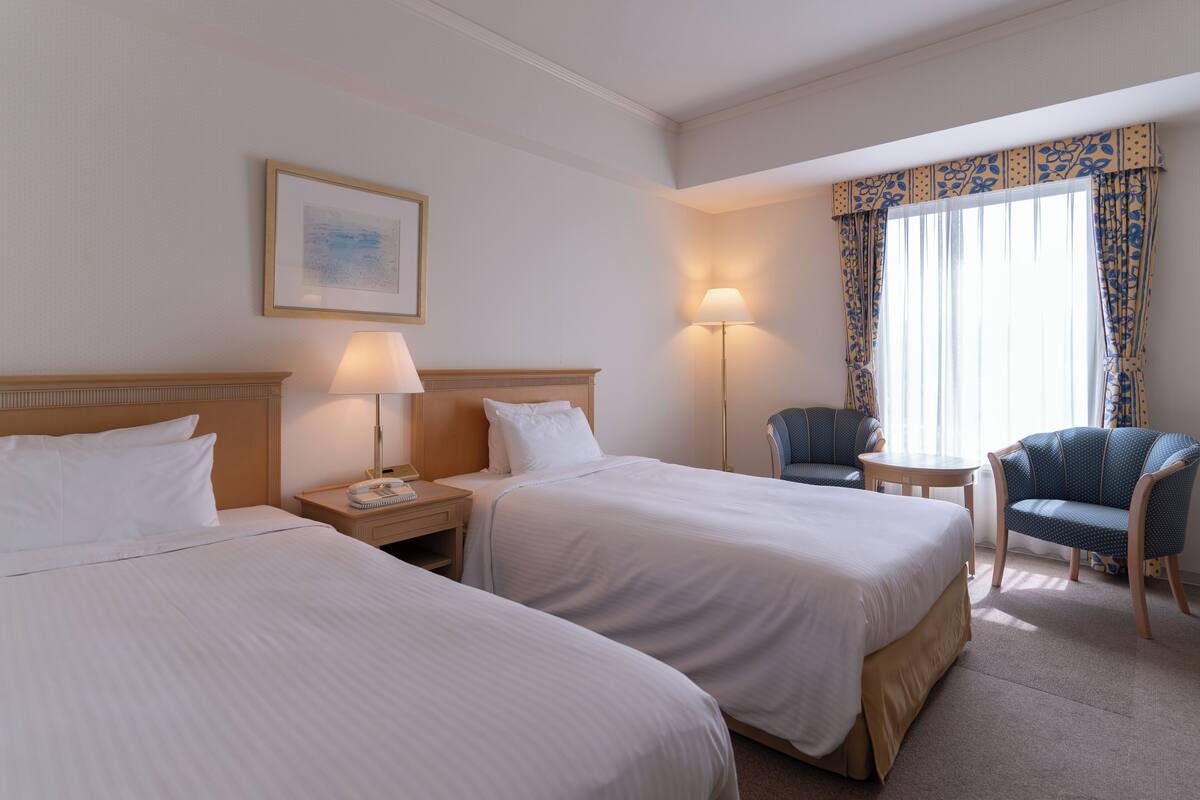 Quarto de hotel com duas camas, uma mesa de cabeceira com abajur, uma cadeira e uma janela com cortinas.