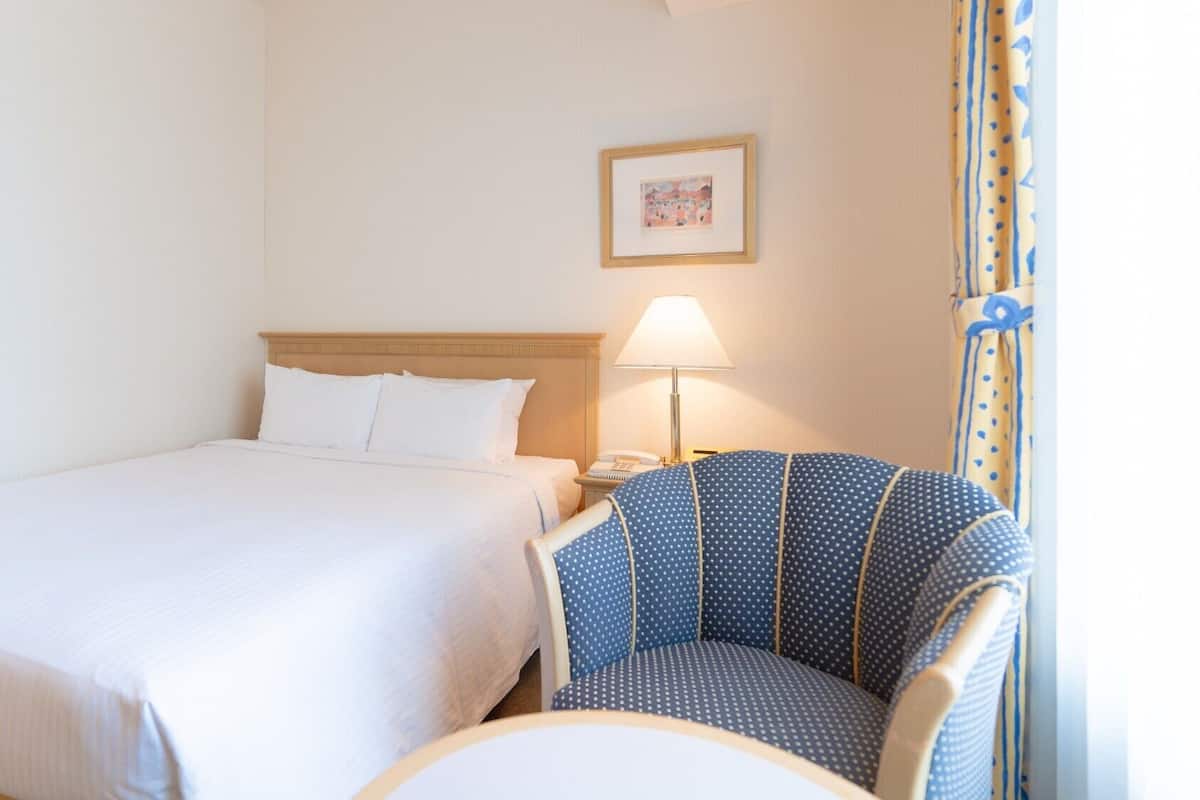 Hotellihuone, jossa on sänky, tuoli, yöpöytä, lamppu ja seinällä oleva kehystetty kuva.