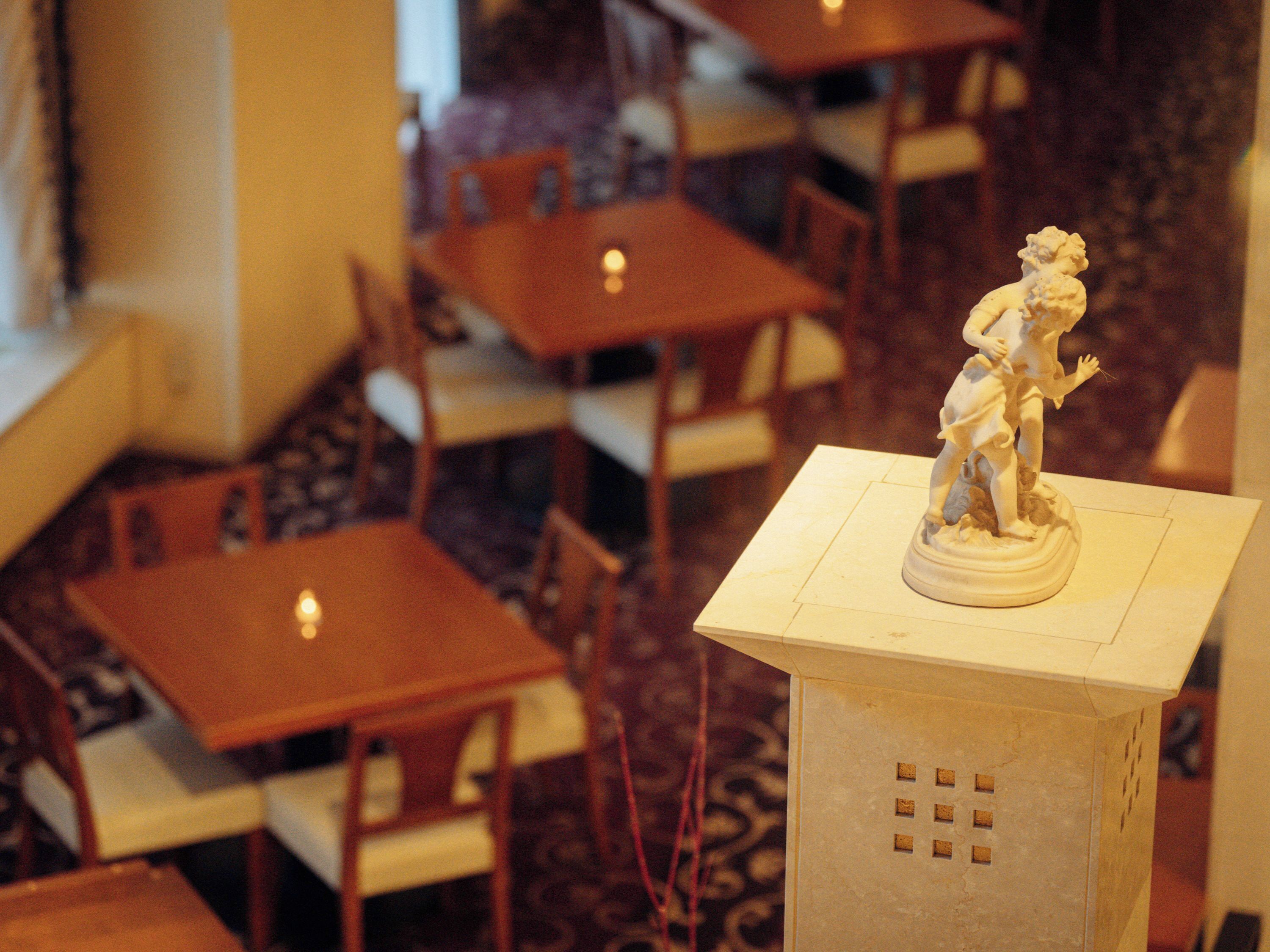 Une statue posée sur un piédestal, dans une pièce où se trouvent plusieurs tables et chaises en bois.