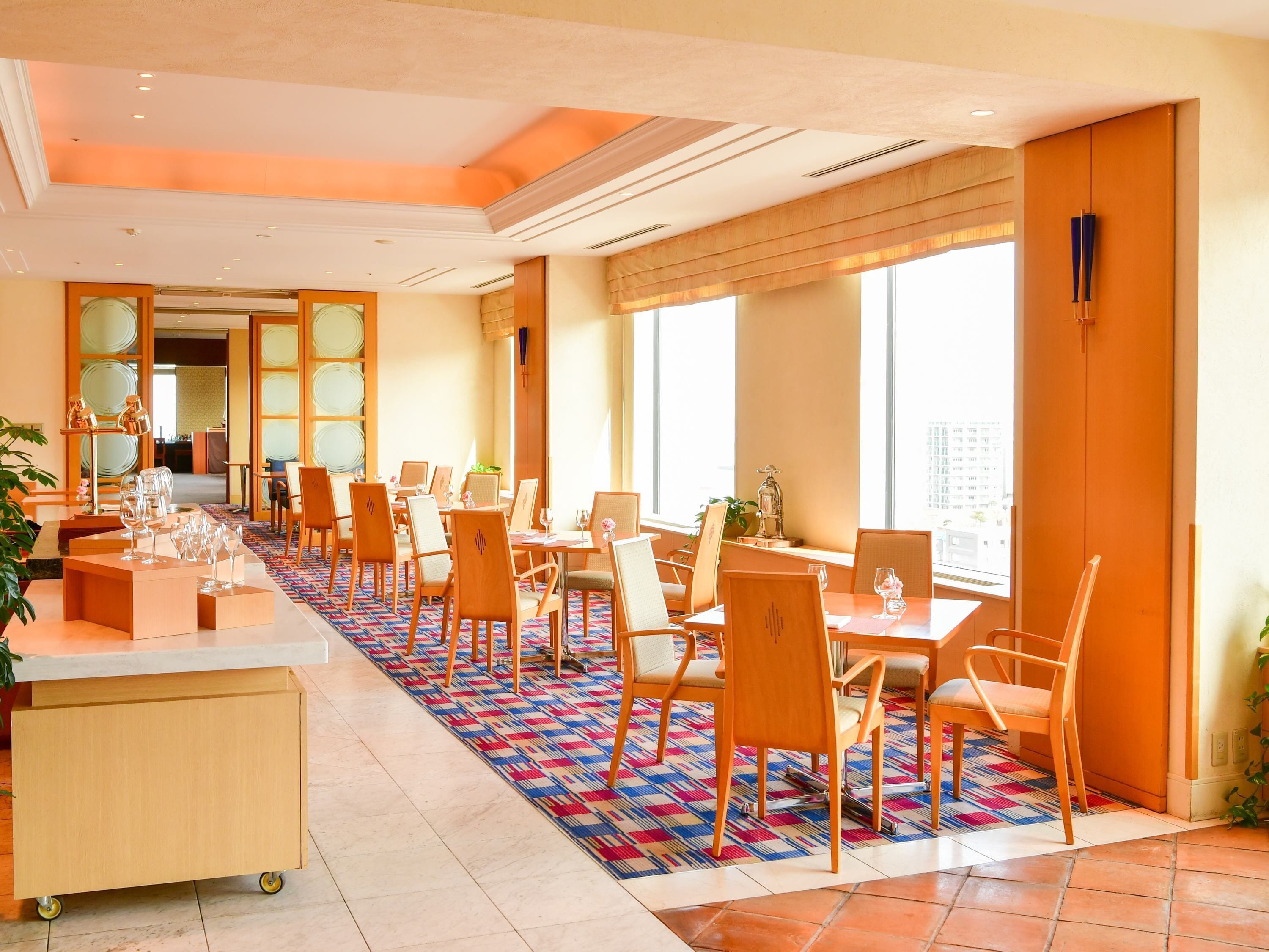 A hotel dining area with multiple tables and chairs, a patterned carpet, and large windows.