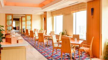 A hotel dining area with multiple tables and chairs, a patterned carpet, and large windows.