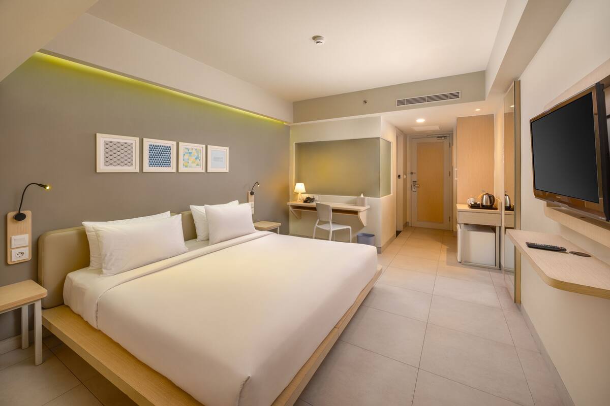 A modern hotel room with a large bed, a desk, a chair, a TV, and artwork on the wall.