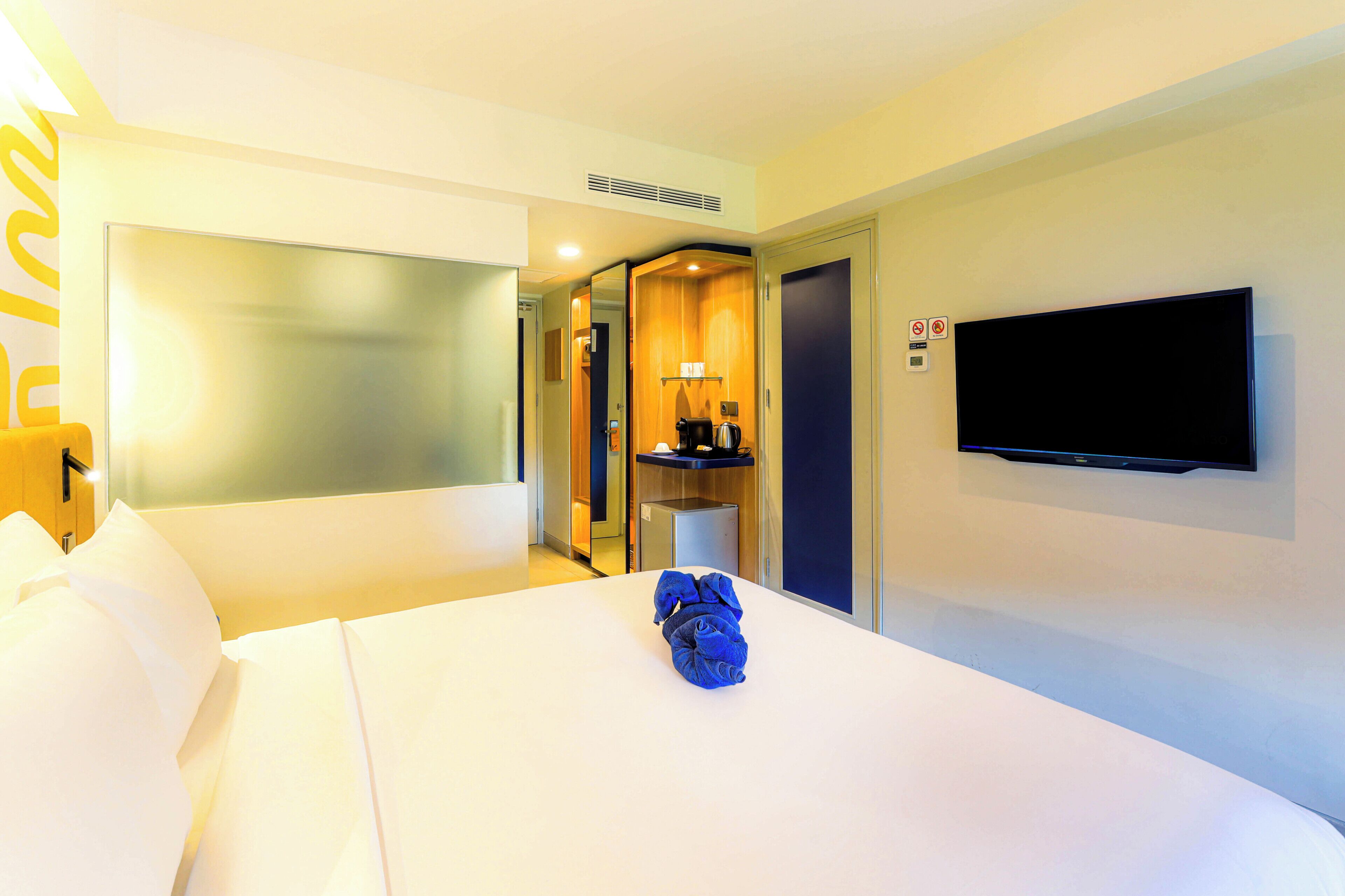 A hotel room with a bed, a flat-screen TV mounted on the wall, a coffee machine, and a door leading to a bathroom.