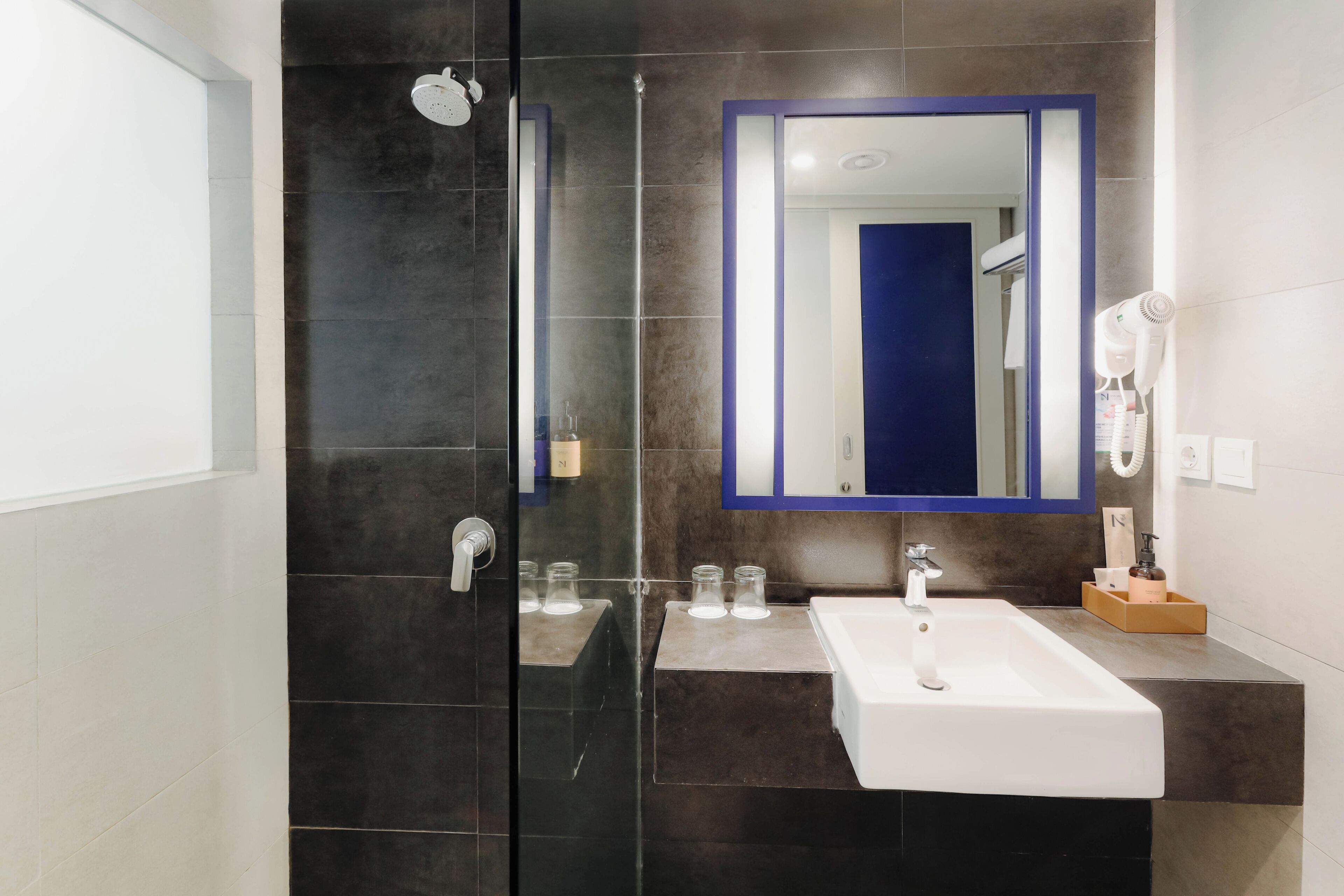 A modern bathroom with a glass-enclosed shower, a white sink, a large mirror, and a hairdryer on the wall.