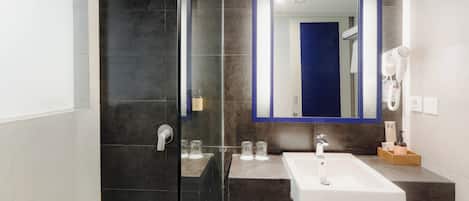 A modern bathroom with a glass-enclosed shower, a white sink, a large mirror, and a hairdryer on the wall.