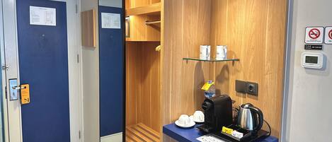A hotel room with a blue door, a wooden panel wall, a coffee station with a kettle, and a digital clock.