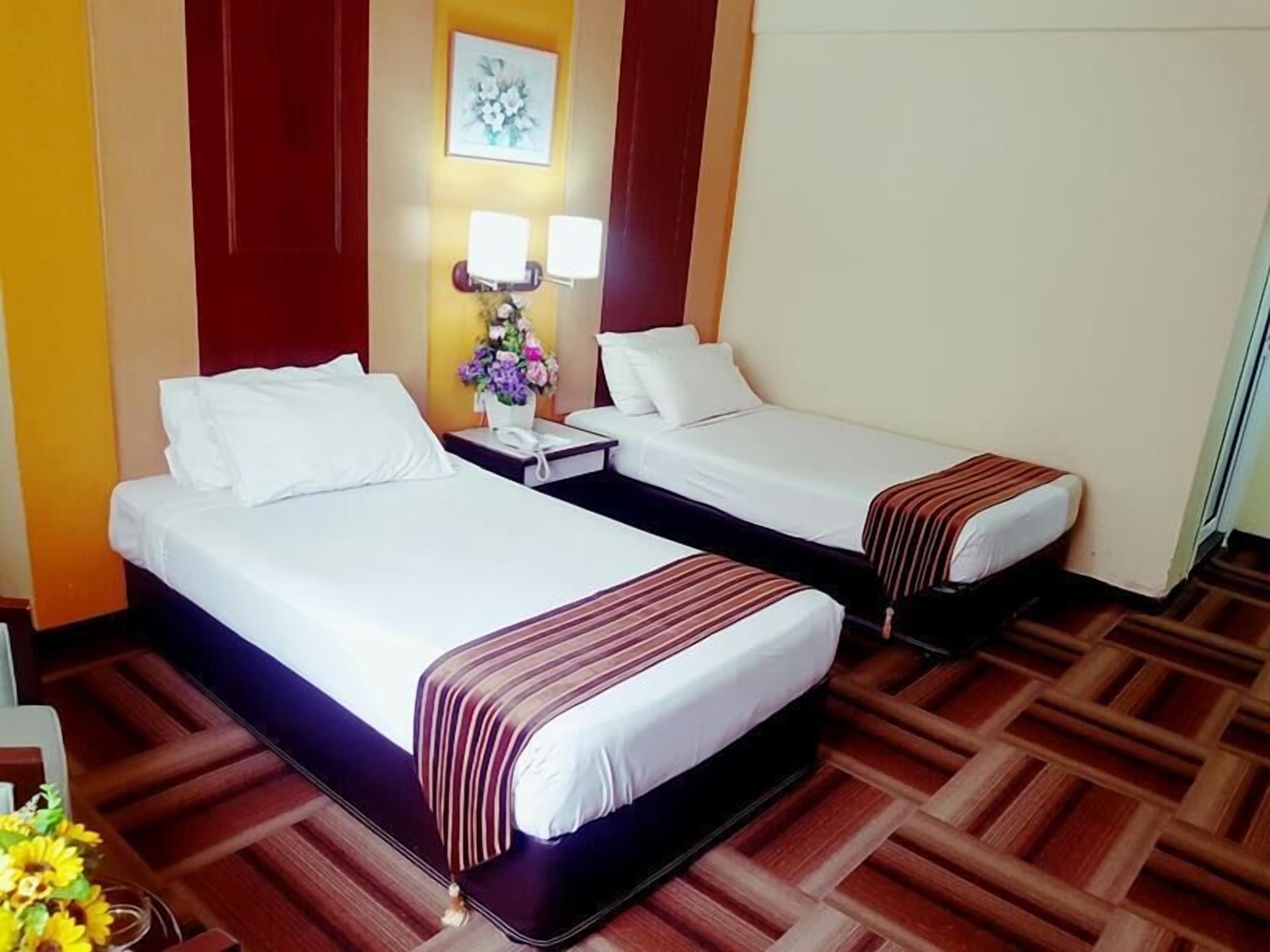 A hotel room with two beds, a nightstand with a lamp, a vase with flowers, and a wooden floor pattern.