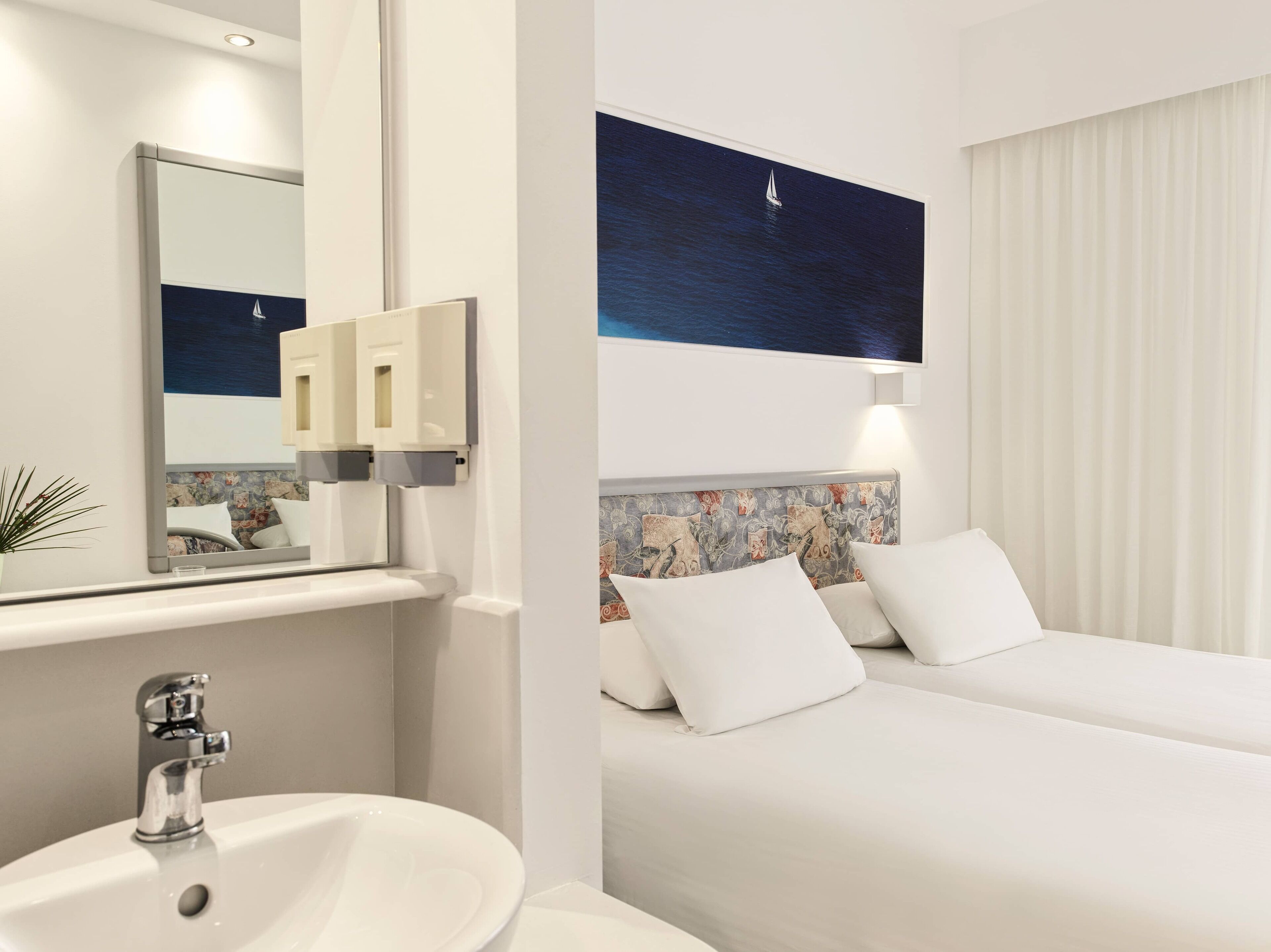 Habitación de hotel con una cama grande, una mesita de noche, un espejo, un lavabo y una lámpara fijada a la pared.