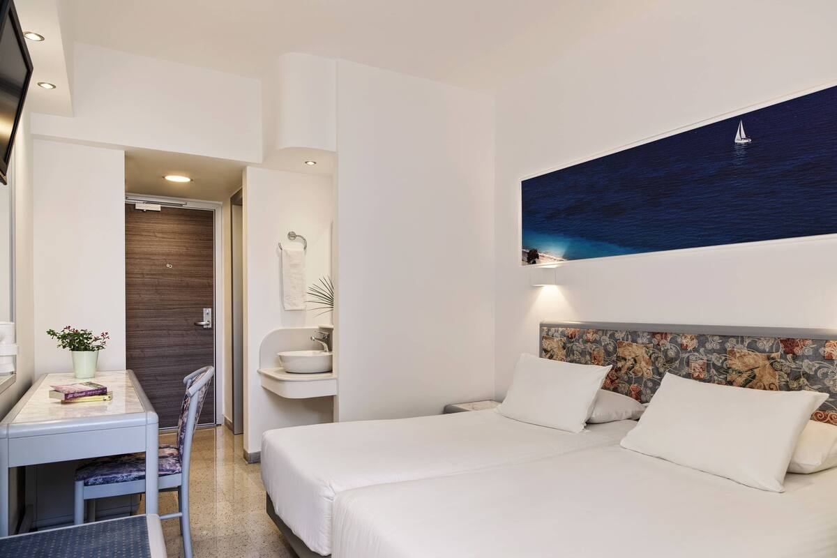 A hotel room with a bed, a desk, and a chair. There is a painting on the wall and a window with a view of the sea.