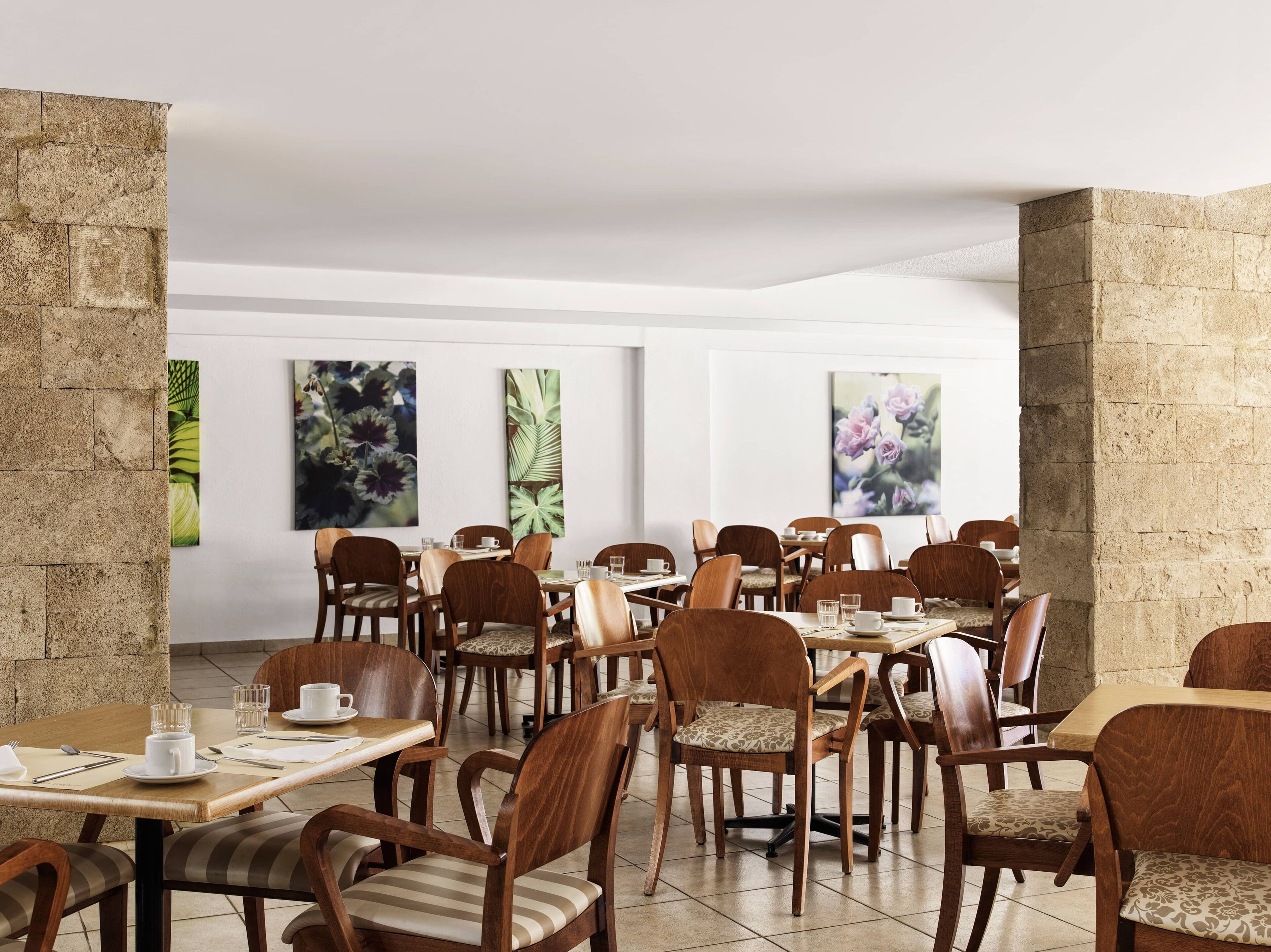 A spacious dining area with wooden tables and chairs, white walls, and large floral artwork.