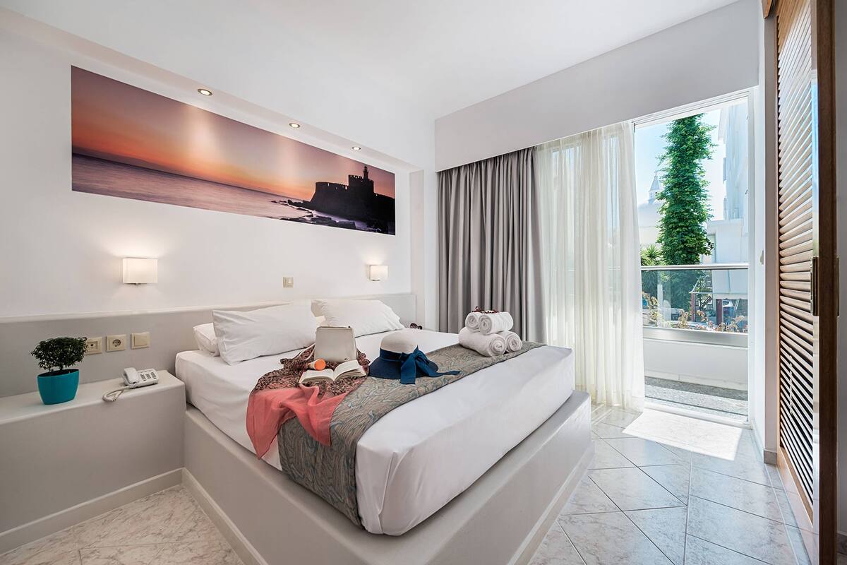 A modern hotel room with a large bed, a painting of a sunset, a balcony with curtains, and a small plant on the nightstand.