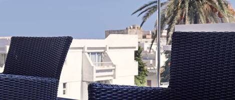 A balcony with wicker chairs and a small table, overlooking a cityscape and palm trees.