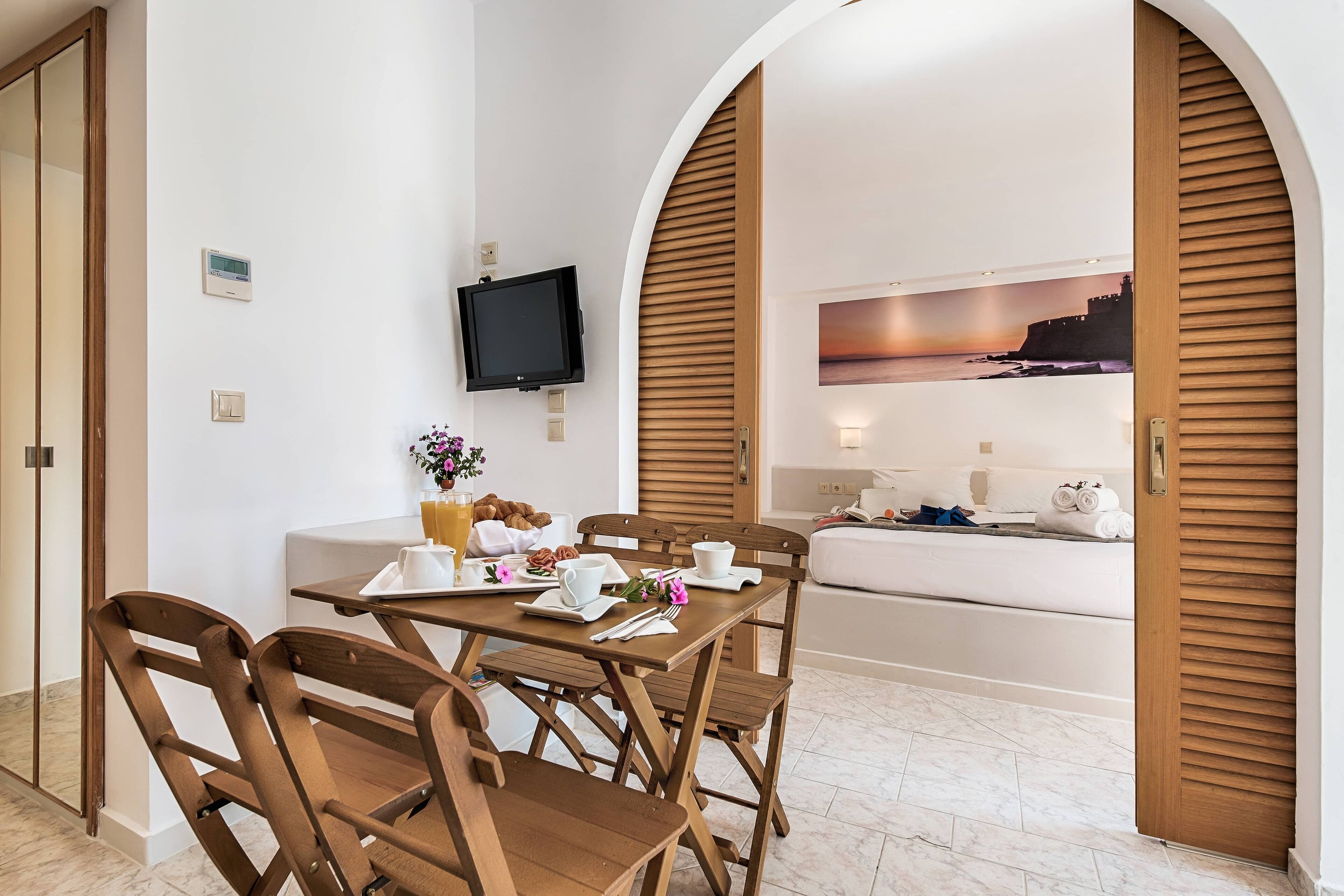 Un coin repas avec une table dressée pour deux personnes, un téléviseur fixé au mur et un tableau représentant un coucher de soleil sur un paysage côtier.