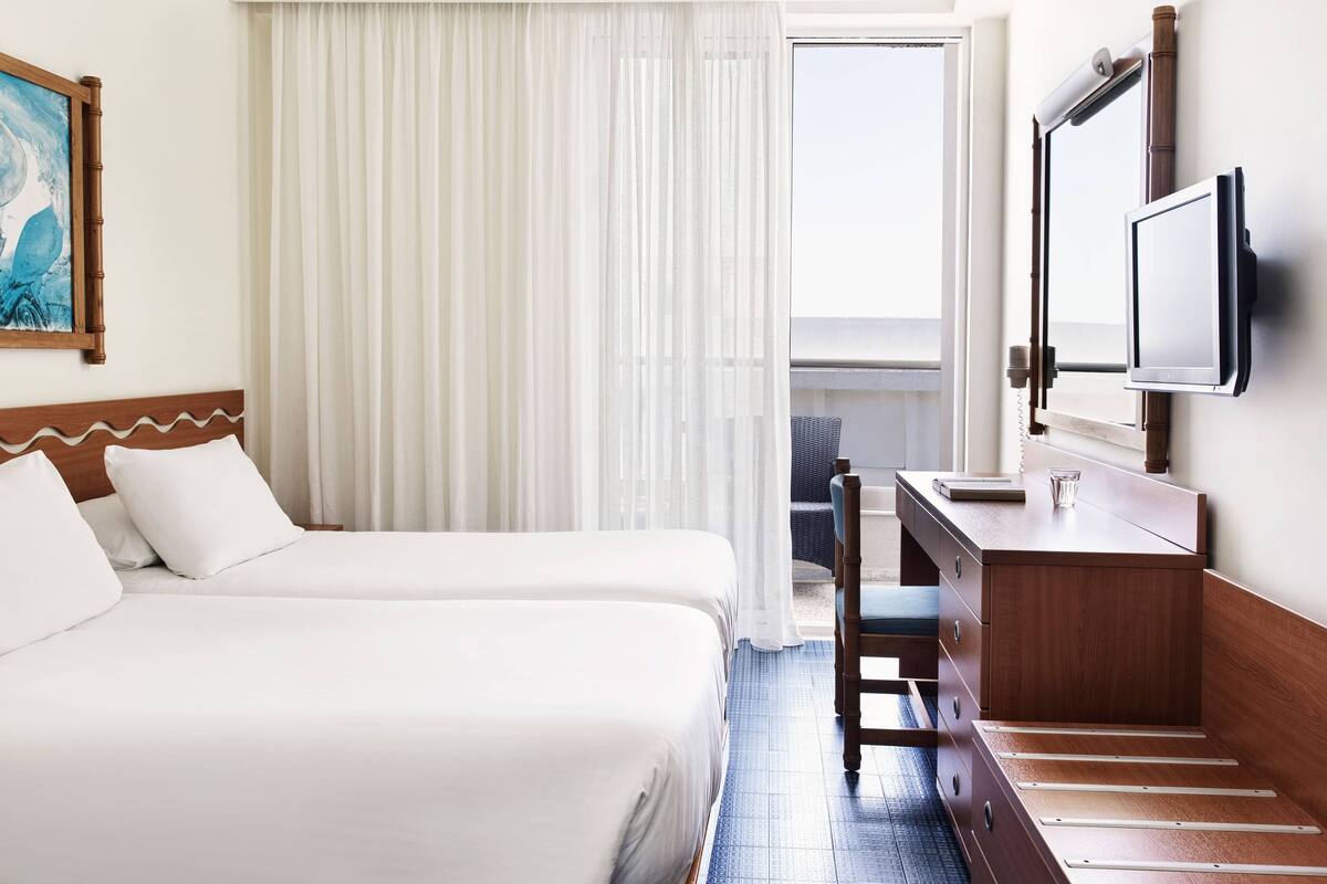 A hotel room with two beds, a desk, a TV, and a large window with curtains.