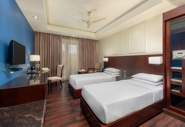A hotel room with two beds, a desk, a chair, a TV, and a ceiling fan. - The Acacia Hotel & Spa Goa (Candolim)