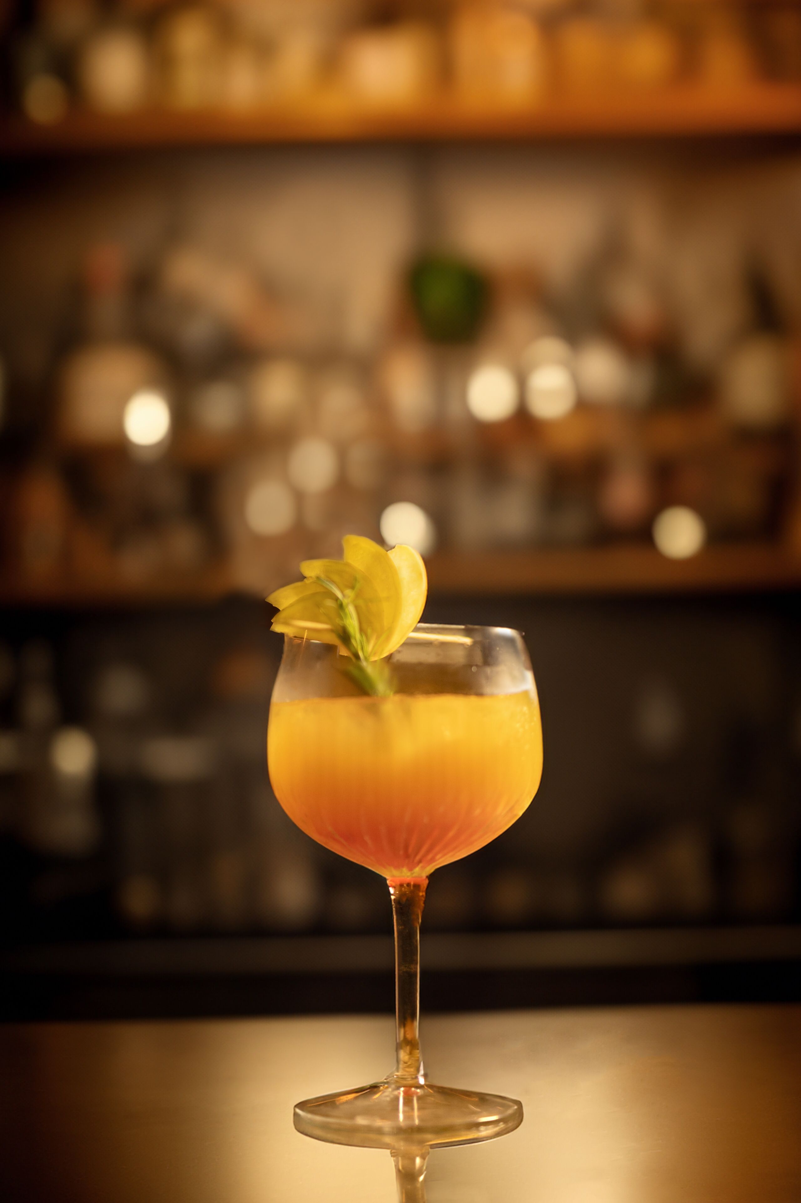 Un vaso de cóctel con una bebida amarillo-anaranjada y una guarnición sobre una barra.