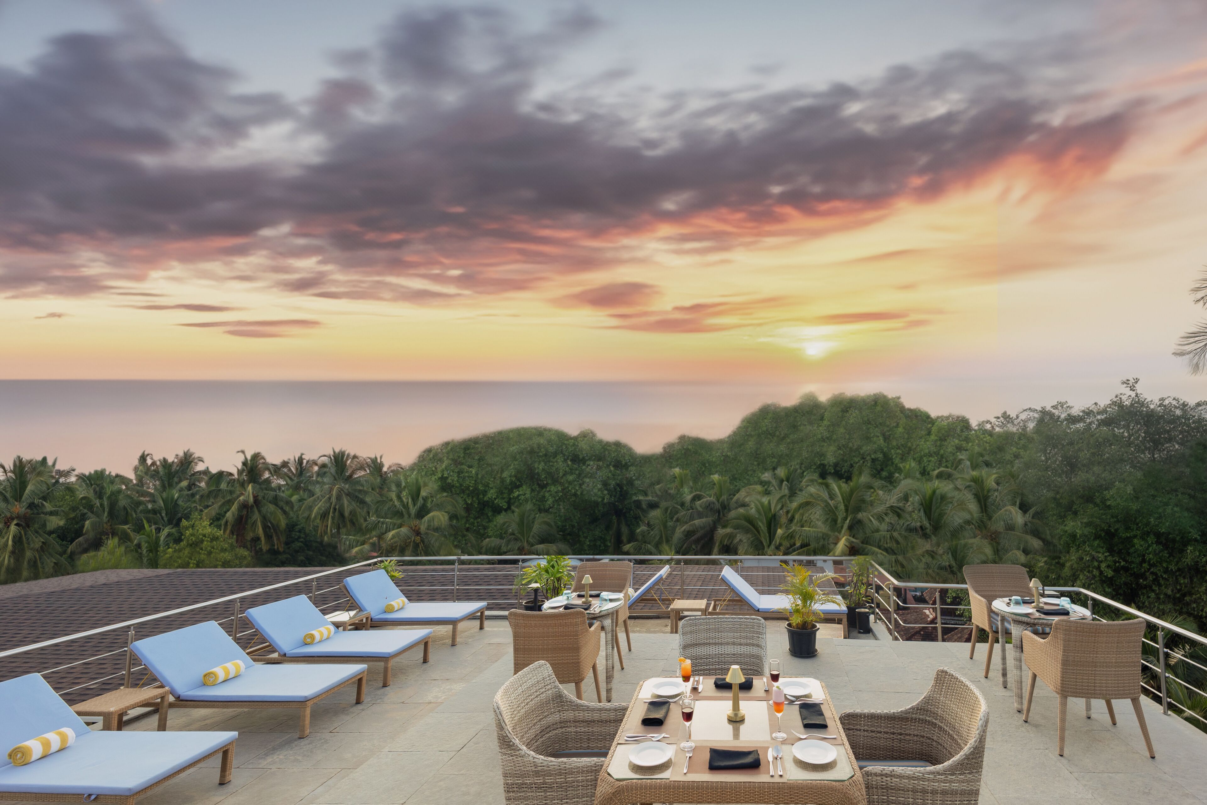 A rooftop terrace with lounge chairs and tables, overlooking a sunset over the ocean.