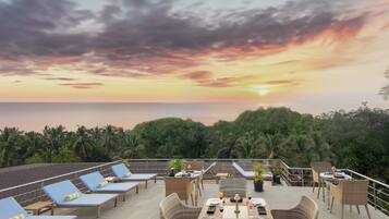 A rooftop terrace with lounge chairs and tables, overlooking a sunset over the ocean.