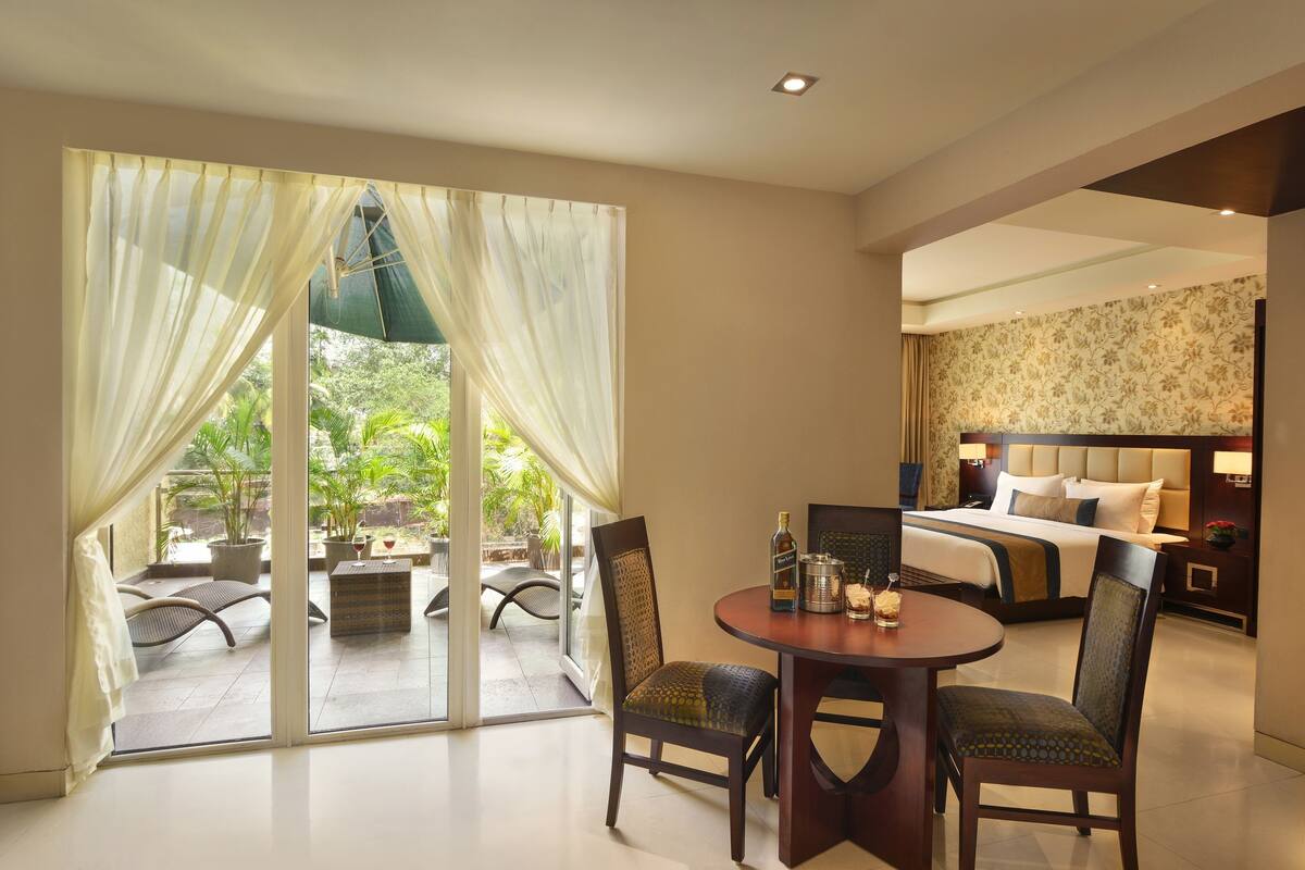 Ein Hotelzimmer mit einer großen Glasschiebetür, die auf eine Terrasse führt, einem runden Esstisch mit Stühlen, einem Bett mit Kopfteil und einem Nachttisch mit Lampe.