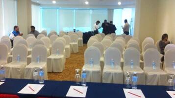 Un salón de conferencias con filas de sillas blancas, mesas con botellas de agua y sillas rojas al frente.