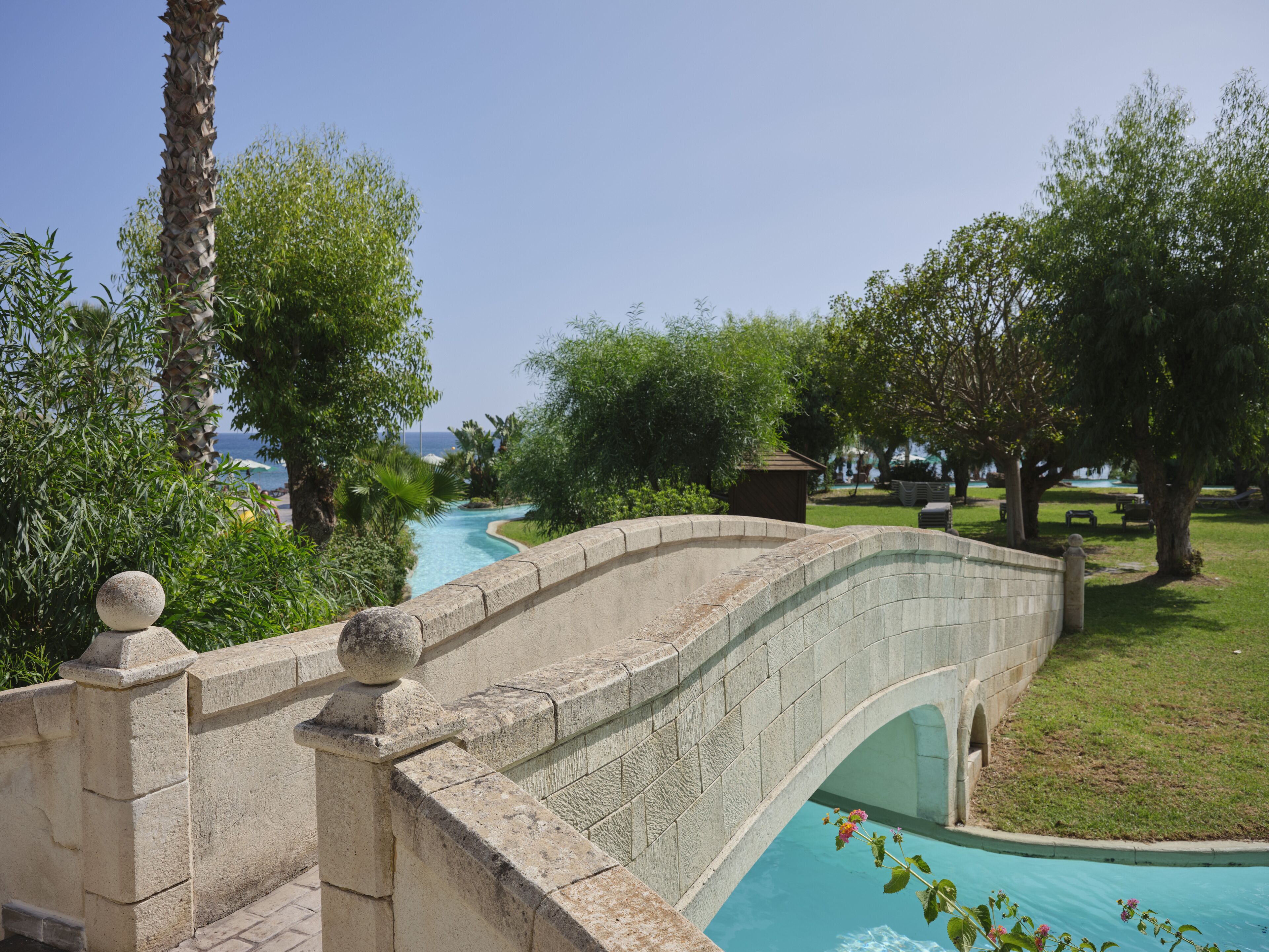 Eine Stebrücke über einem Schwimmbad, umgeben von üppiger Vegetation und Blick aufs Meer.
