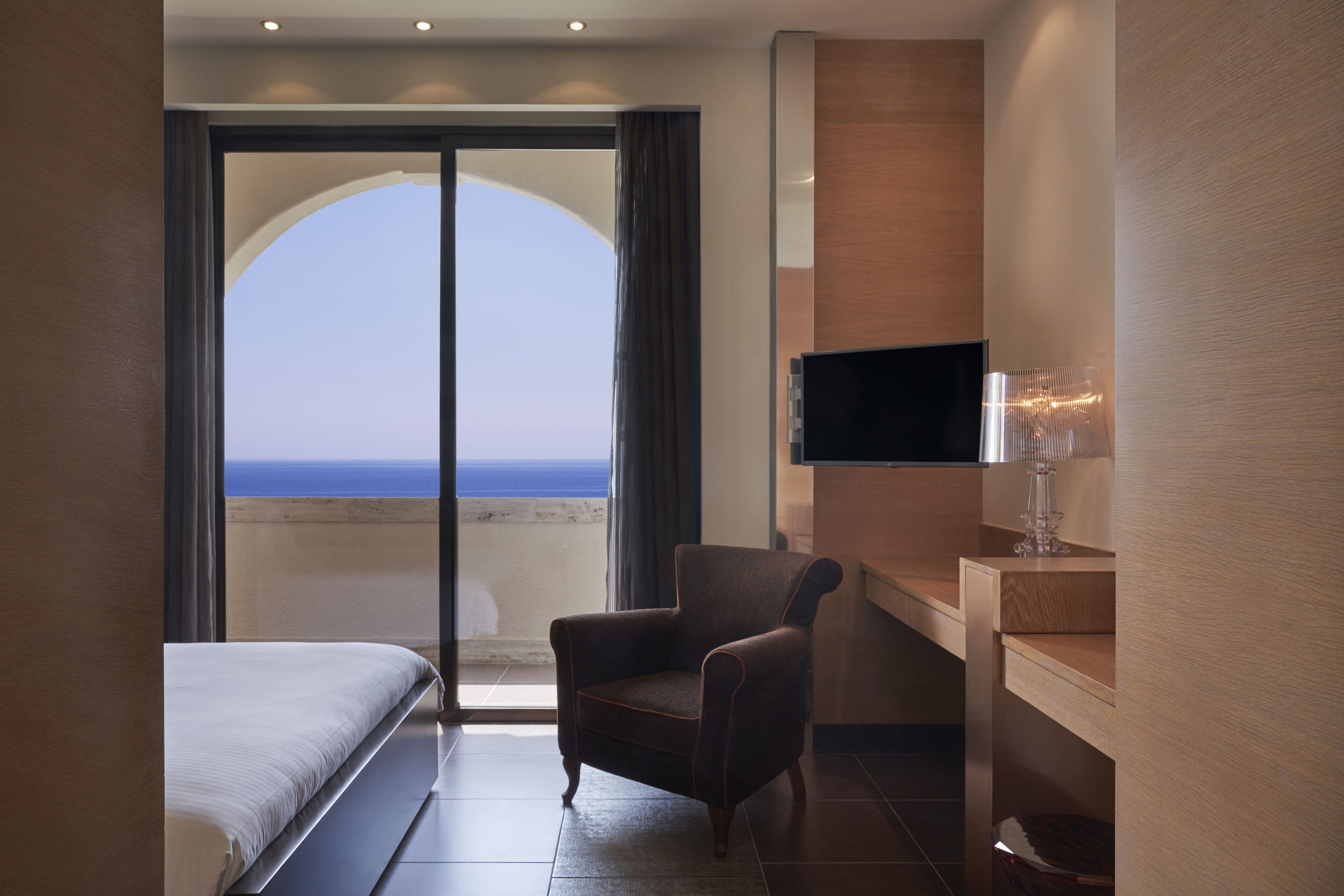 Una habitación de hotel con cama, sillón, televisión y vista al océano a través de un ventanal grande.