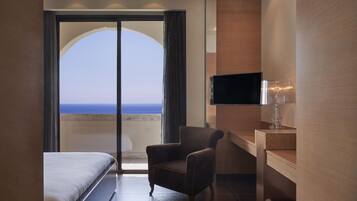 Una habitación de hotel con cama, sillón, televisión y vista al océano a través de un ventanal grande.