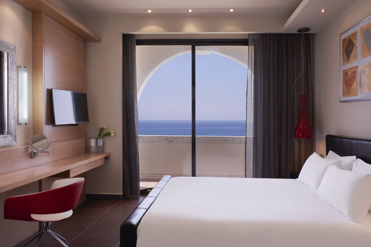 Habitación de hotel con una cama grande, un escritorio con una silla, un televisor y vistas al océano a través de una puerta corrediza.