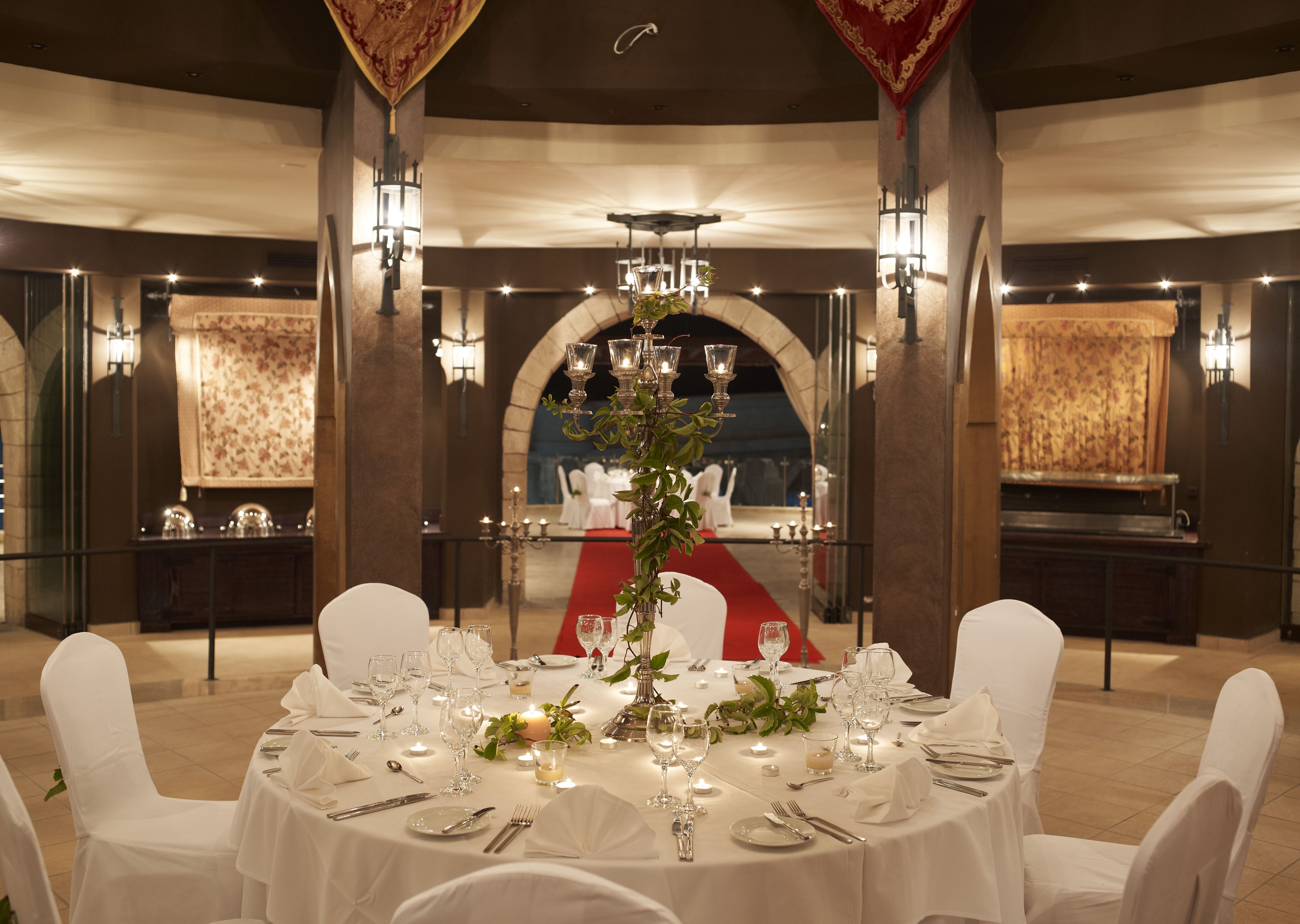 Un comedor formal con mesas redondas dispuestas para una comida, sillas blancas y una alfombra roja que lleva a una zona de estar.