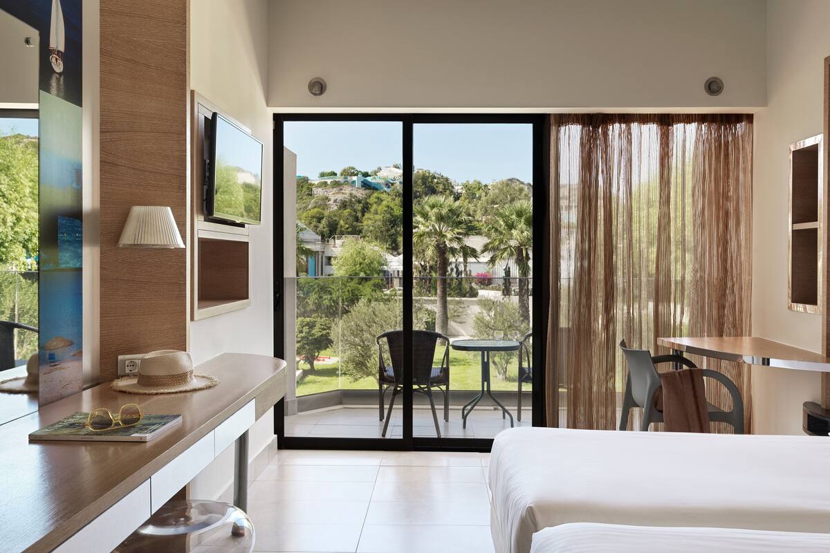 Habitación de hotel con una cama, un escritorio con lámpara, una silla y vistas a un balcón con mesa y sillas.
