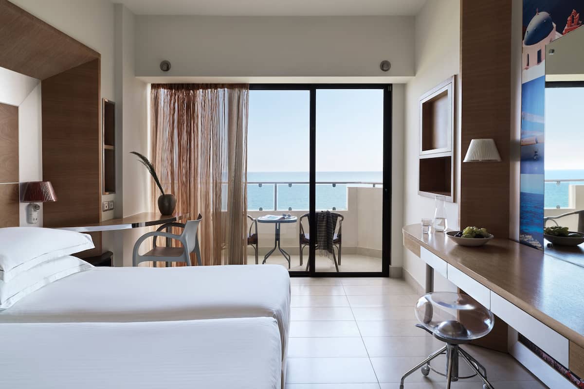 Habitación de hotel con cama, escritorio, silla, mesita, televisor y balcón con vista al mar.