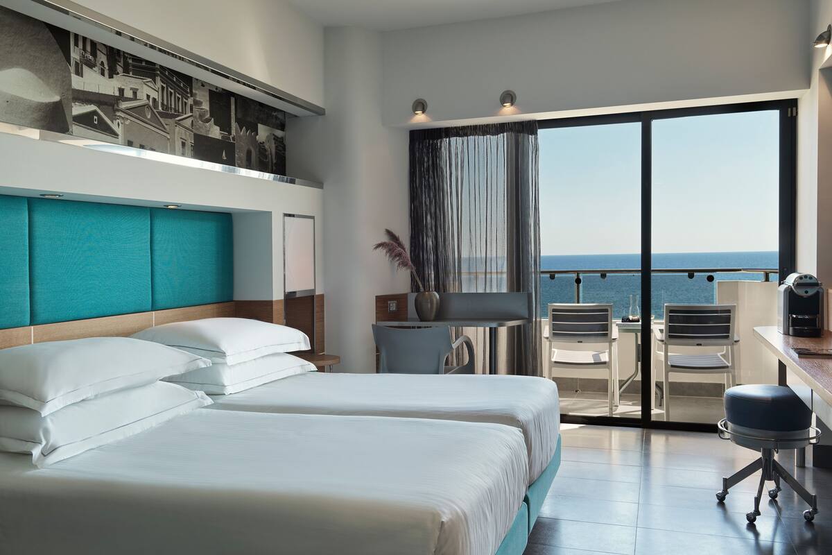 Habitación de hotel con dos camas, un escritorio con silla, un ventanal con vista al mar y un balcón con mesa y sillas.