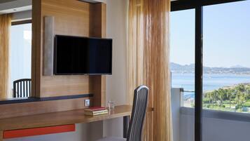 Habitación de hotel moderna con un televisor grande montado en un panel de madera, una pequeña mesa con una silla, un vaso de agua y vista al mar a través de un ventanal.